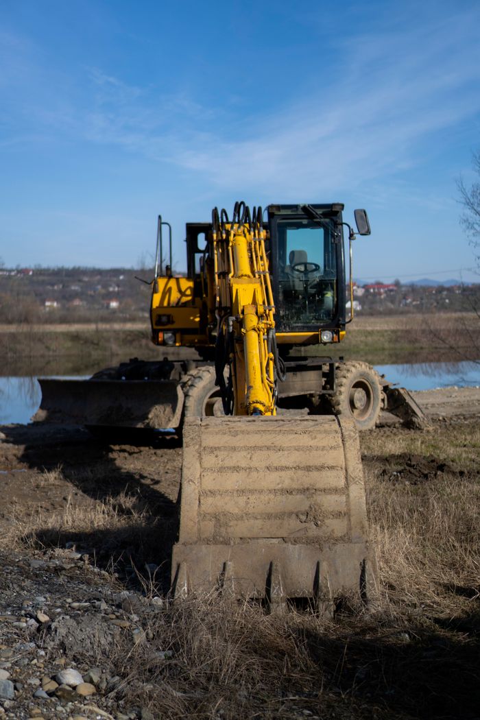 A yellow excavator at work on a riverbank construction site under a clear blue sky.
