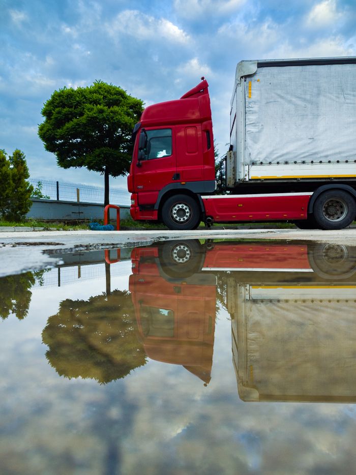 A parked red truck with a tarp-covered cargo is reflected in a puddle on an urban street, under a cloudy sky.