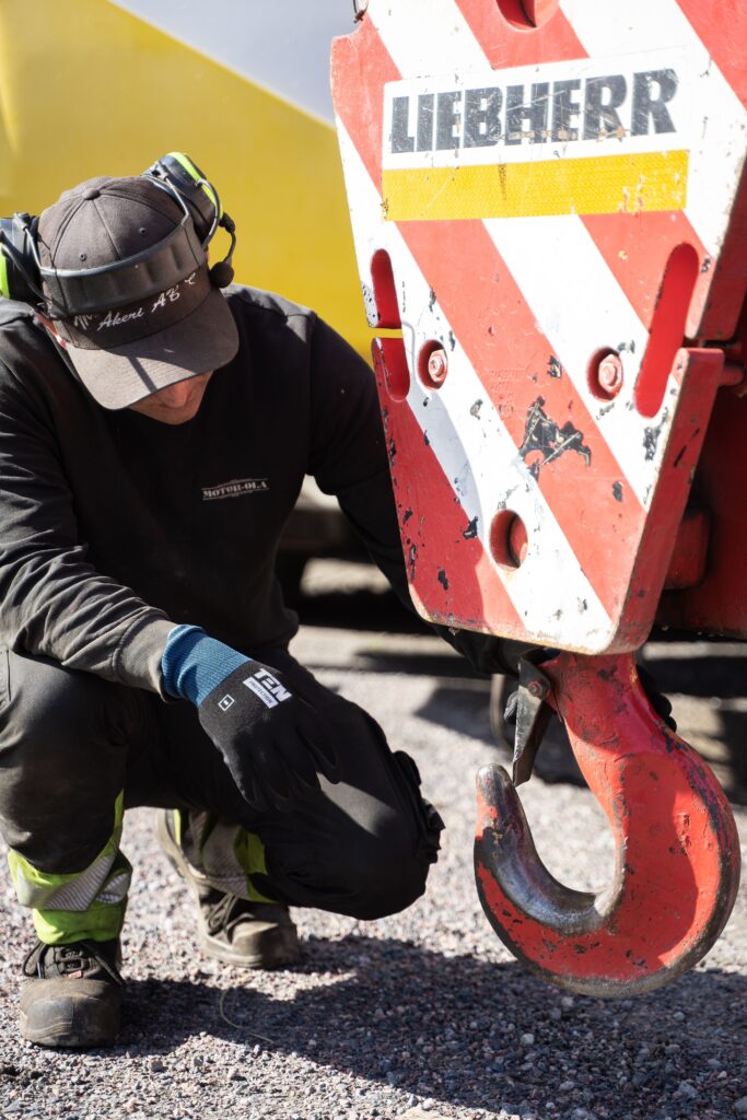 A construction worker crouches to inspect a large machinery hook outdoors, wearing cap and headphones.
