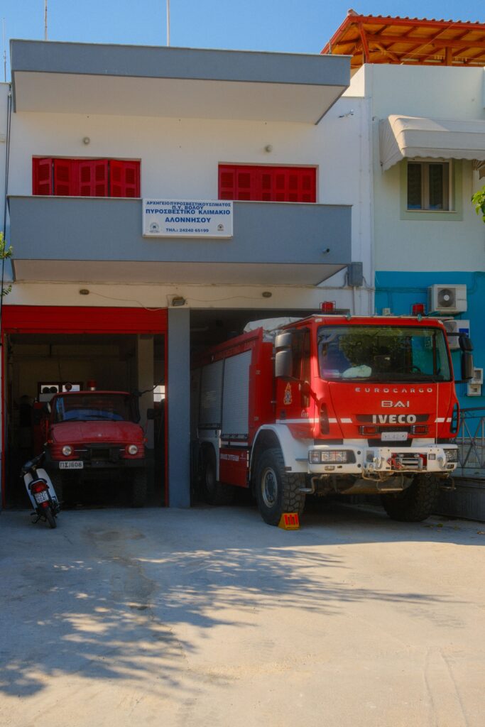 Exterior view of a Greek fire station with parked red fire trucks and motorcycles.