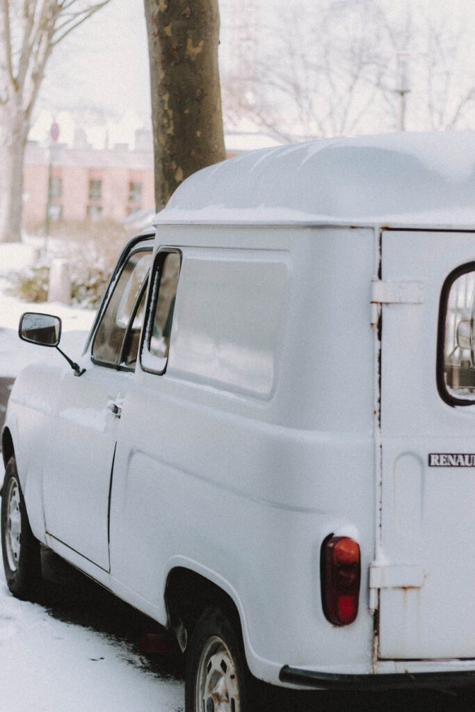 White old timer automobile with rear bumper parked on snowy path against residential house in town on cold winter day
